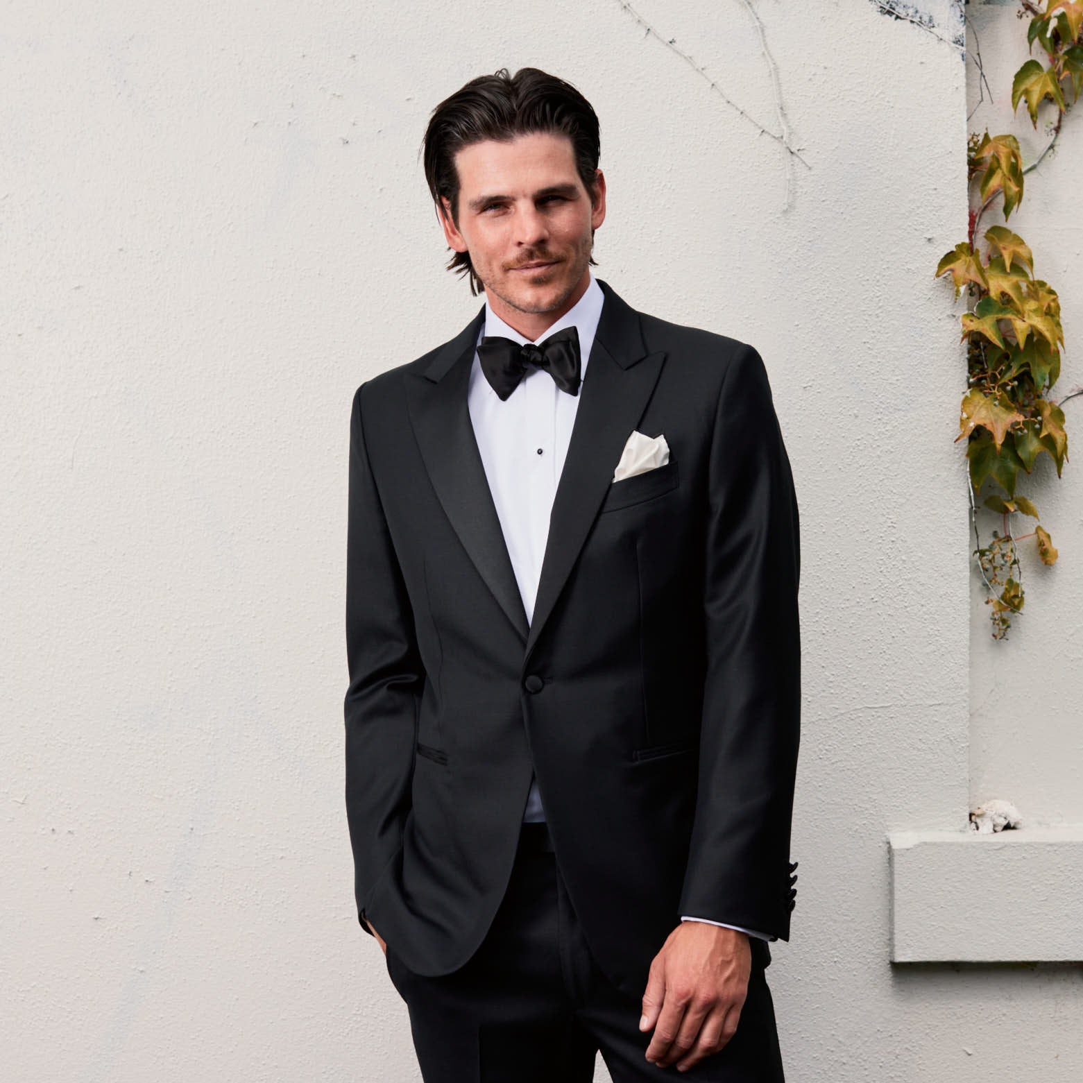 Man in a black tuxedo standing against a white wall with decorative plants.